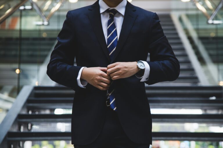 person in a dark business suit adjusting their jacket while standing in front of a staircase, wearing a striped tie and wristwatch, head not visible