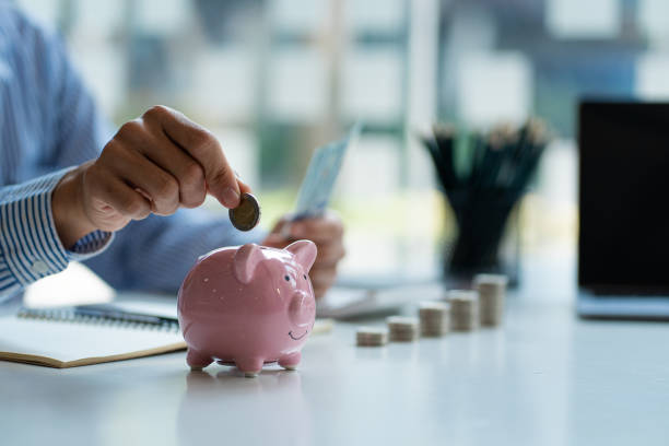 person placing a coin into a pink piggy bank on a desk with stacked coins, notebook, and laptop in the background—symbolising savings and one of the best money advice principles for building financial security