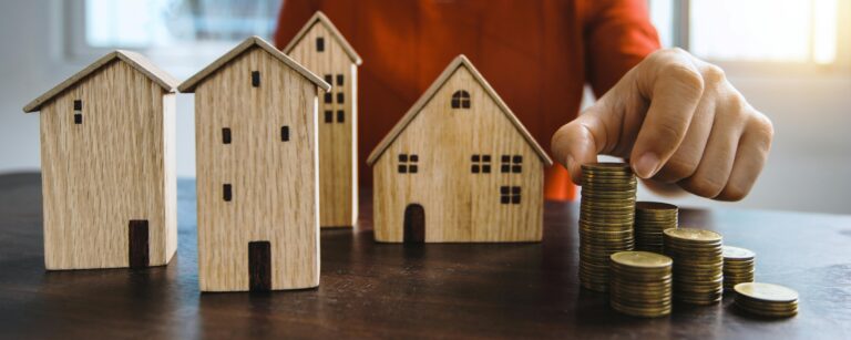 hand stacking gold coins next to small wooden house models representing property investment and building wealth